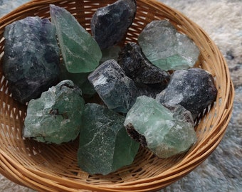 May include: A wicker basket containing raw fluorite crystals. The crystals are various shades of green and purple, with a natural, unpolished look. The basket is made of light brown woven material.