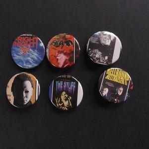 May include: Six round pinback buttons featuring horror movie posters. The buttons display images and titles from films like "Night of the Demons", "The Stuff", and "Hellbound: Hellraiser II". The buttons are arranged on a black surface.