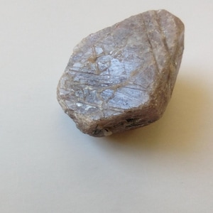 Puede incluir: Una gema en bruto, de forma irregular, de color marrón claro, con una superficie texturizada. La piedra tiene un aspecto ligeramente translúcido y muestra sutiles variaciones de color y tono. La forma general es redondeada con algunos bordes angulares.