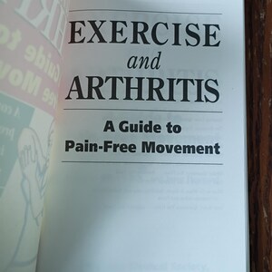 May include: An open book displaying the title "EXERCISE and ARTHRITIS" in bold black text. The subtitle reads "A Guide to Pain-Free Movement." The pages are white, and the background is a wooden surface.