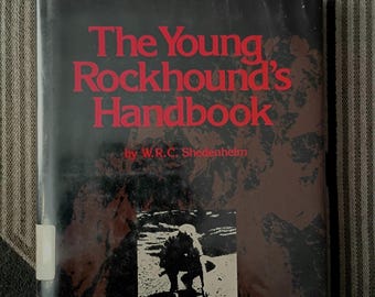 May include: A hardback book titled The Young Rockhound's Handbook by W.R.C. Shedenhelm. The cover is black with dark brown textured patterns and features bold red text for the title. A small square black and white photograph shows a person.