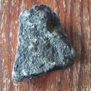 Può includere: Una roccia grigio scuro con una superficie ruvida e strutturata. La roccia ha una forma leggermente irregolare ed è posta su uno sfondo di legno con un tono marrone caldo. Il colore della roccia varia dal grigio scuro al grigio più chiaro.