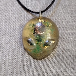 May include: A pendant necklace featuring a resin alien head design. The pendant is a translucent yellow-green with green and gold glitter accents. The alien has two large, oval eyes and a circular detail. The necklace has a black cord.