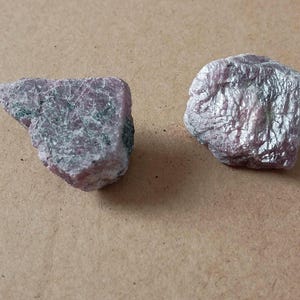 May include: Two rough-cut, pinkish-purple gemstones with a crystalline structure. The stones have a slightly rough texture and appear to have some green inclusions. The background is a plain, light brown surface.