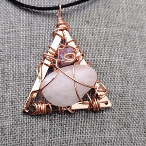 May include: A copper-colored wire-wrapped pendant necklace with a triangular frame. The pendant features a light pink oval stone and a smaller purple stone. The necklace has a black cord.