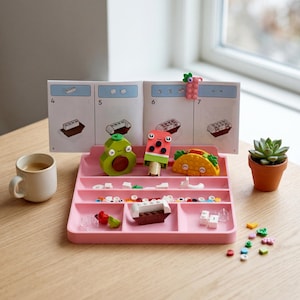 May include: A pink tray with compartments holds colourful Lego food creations: an avocado, watermelon ice lolly, and taco. Building instructions are in the background. A small succulent in a terracotta pot and a cup of coffee are also present.