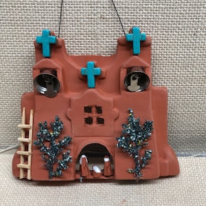 May include: A terracotta-colored ceramic ornament depicting a church with a ladder, two small trees, and three figures in the doorway. The church has three turquoise crosses on the roof and two silver bells.