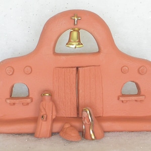 May include: A terracotta nativity scene with a small bell in the archway, a gold cross, and three figures: a wise man, Mary, and the baby Jesus.