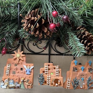 May include: Three terracotta-colored clay ornaments featuring Southwestern-style adobe buildings. One depicts a nativity scene with a star, another shows a multi-story structure with ladders, and the third features blue crosses and small metallic bells.