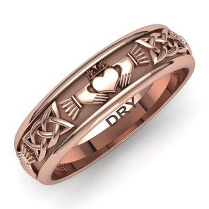May include: A rose gold band ring with a Celtic knot design and a Claddagh symbol featuring two hands holding a heart with a crown on top. The inscription "DRY" is visible on the inside of the band.