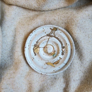 Op de afbeelding: Een witte keramische sieradenschaal met bruine spikkels bevat een gouden ring met een groene steen, een gouden ketting en een goudkleurige paperclip in de vorm van een veer. De schaal staat op een beige, getextureerd oppervlak. Een kleine gouden hanger is ook zichtbaar.