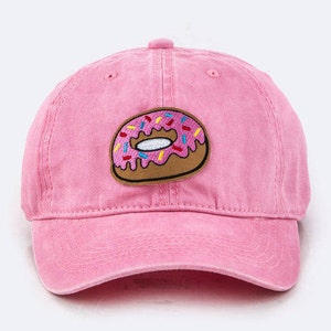 May include: Pink baseball cap with a donut patch. The cap is a solid pink color with a curved brim and a stitched donut patch on the front. The donut patch has brown frosting, colorful sprinkles, and a white center.