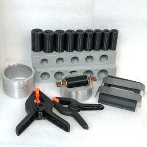 May include: A collection of tools, including black and orange clamps, a gray holder with black cylinders, and a silver metal cylinder. The tools are arranged on a white surface. The black cylinders are labeled with numbers.
