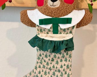 May include: A Christmas stocking shaped like a teddy bear, with a brown plush head and a green and beige patterned body. The stocking has a white collar, a green bow, and a green ruffle. The name "Mike" is written on the toe.