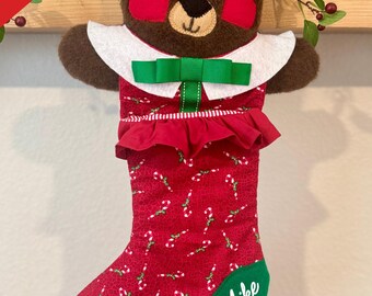 May include: A Christmas stocking shaped like a teddy bear, with a red body patterned with candy canes. The bear has a brown head, tan ears, and red heart-shaped cheeks. A green bow adorns the white collar. The name "Mike" is embroidered on the green toe.