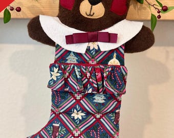 May include: A Christmas stocking shaped like a teddy bear, with a brown head and a patterned body. The stocking features a white collar, a burgundy bow, and a patterned fabric with Christmas motifs. The name "Mike" is written on the toe.