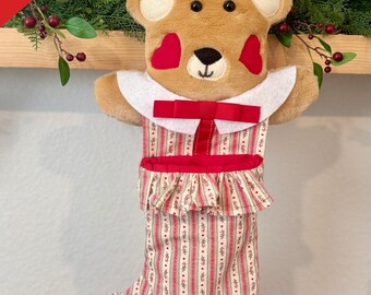 May include: A Christmas stocking shaped like a teddy bear, with a red and white striped pattern. The bear has a white collar, red bow, and heart-shaped cheeks. The name "Mike" is embroidered on the cuff. The text "Custom Name Available" is on a red banner.