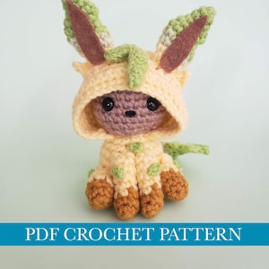 Può includere: Bambola amigurumi all'uncinetto in un completo con cappuccio giallo chiaro con dettagli verdi e marroni. La bambola ha una testa rotonda, occhi neri e un naso marrone. Il testo "PDF CROCHET PATTERN" è in basso.