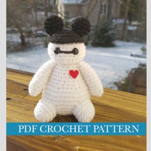May include: White crocheted amigurumi toy with black Mickey Mouse ears, black eyes, and a red heart on its chest.  The toy is sitting on a wooden surface.  PDF CROCHET PATTERN.