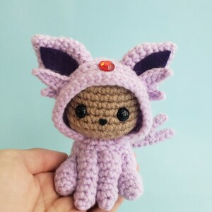 May include: A small crocheted plush toy wearing a purple and black hooded costume with a red gem on the top of the hood. The toy has brown yarn for the face and black eyes.