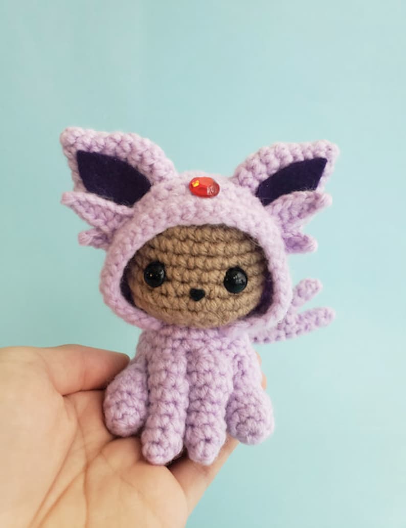 May include: A small crocheted plush toy wearing a purple and black hooded costume with a red gem on the top of the hood. The toy has brown yarn for the face and black eyes.