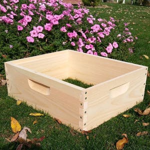 May include: A wooden beehive box with a handle on the front. The box is made of natural wood and has a light brown color. The box is sitting on a bed of green grass with some fallen leaves.