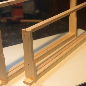 May include: Two unfinished wooden bee hive frames. The frames are rectangular with horizontal and vertical wooden slats. The wood is light-colored and appears to be new. The frames are sitting on a white surface.