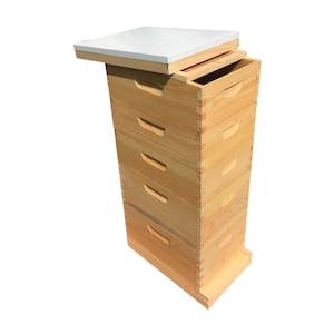 May include: A wooden beehive with a white lid, consisting of five stacked boxes with a base. The boxes have a simple design with a small handle on each side.