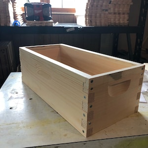 May include: A wooden box with a handle on the side. The box is made of light-colored wood and has a natural finish. The box is sitting on a white surface.