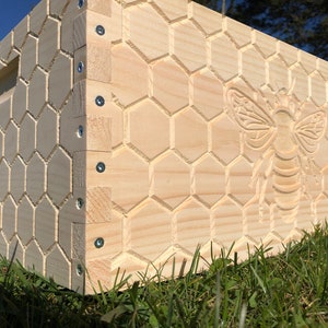 May include: A wooden beehive box with a carved honeycomb pattern and a large bee design. The box is made of light-colored wood and has a handle on one side. The bee is intricately carved, adding a decorative element to the functional design.
