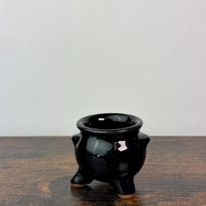 May include: A small, round, black ceramic cauldron with a glossy finish. The vessel features a wide, open top rim, two small side handles, and three short, sturdy legs with exposed brown clay bases, creating a stable, tripod-like structure.