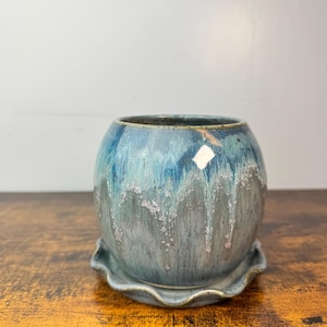 May include: A round ceramic planter with a matching saucer. The planter is a mix of blue and gray with a dripped glaze effect. The saucer has a scalloped edge. The planter sits on a wooden surface.