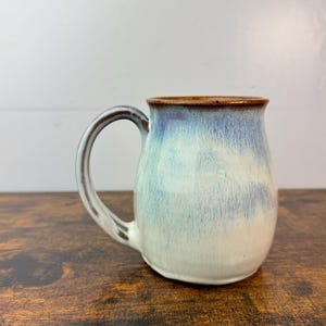 May include: A handmade ceramic mug with a rounded body and a sturdy handle. The mug features a gradient of colors, transitioning from a light blue at the top to a creamy white at the bottom. The rim and handle are a dark brown.