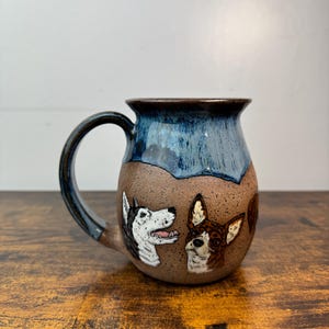 May include: A handmade ceramic mug with a blue and brown glaze. The mug features two dog portraits, one a husky and the other a brown and white dog. The handle is dark blue. The mug sits on a wooden surface.