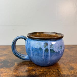 May include: A handmade ceramic mug with a rounded body and a dark brown rim. The mug features a blue glaze with white drips and a matching blue handle. The mug is sitting on a wooden surface.