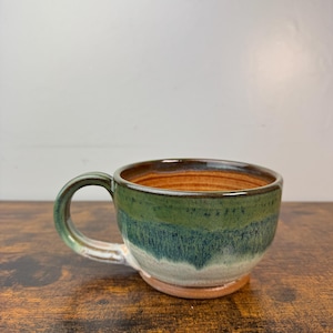 May include: A handmade ceramic mug with a rounded shape and a handle. The mug features a gradient of colors, including green, blue, and brown. The interior is a warm brown color, and the base is a natural clay color.