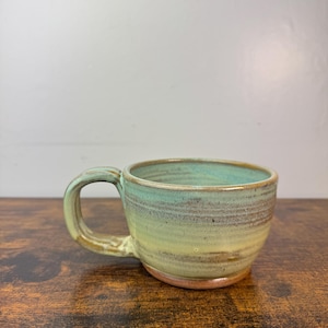 May include: Handmade ceramic mug with a rounded body and a sturdy handle. The mug features a gradient of colors, transitioning from a light green at the top to a yellow-brown at the base. The interior is a light blue-green color.