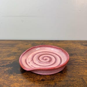 May include: A pink ceramic spoon rest with a spiral design in the center. The spoon rest has a curved shape with a raised edge and a small lip for holding a spoon. The color is a blend of light and dark pink.