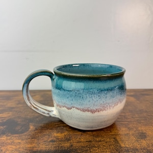 May include: A handmade ceramic mug with a rounded body and a curved handle. The mug features a gradient of colors, transitioning from a deep teal at the rim to white at the base, with a band of reddish-brown. The mug sits on a wooden surface.