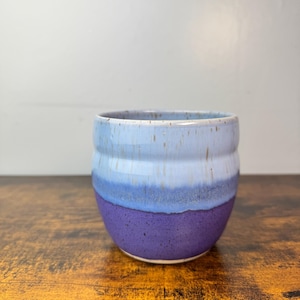 May include: A handmade ceramic cup with a gradient of colors. The cup transitions from a deep purple at the base, through shades of blue, to a light blue rim. The cup is cylindrical with a slightly textured surface.