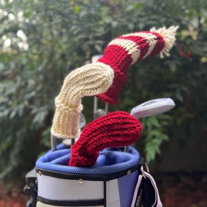 May include: A golf bag with three knitted club covers. One cover is cream-colored, and two are red with white stripes. The golf bag is blue, white, and lavender with a pink shoulder strap. The background is a blurred green.