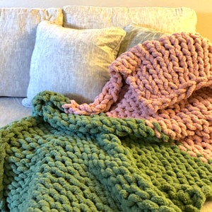 May include: A cozy, hand-knitted blanket in shades of green and pink, draped over a couch. The chunky knit texture is visible, with a soft, inviting appearance. The blanket is paired with light-colored pillows.