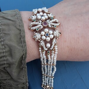 K&ouml;nnte beinhalten: Ein goldenes Armband mit einem mehrreihigen Perlendesign. Die Mitte des Armbands zeigt einen Cluster aus Perlen und bunten Edelsteinen.