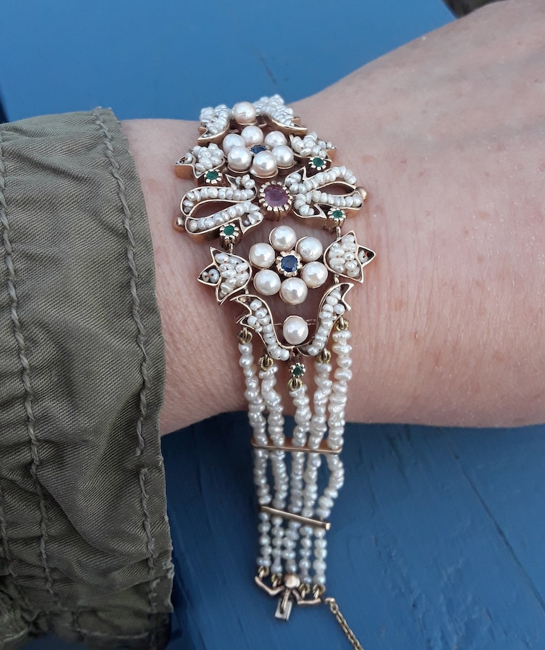 K&ouml;nnte beinhalten: Ein goldenes Armband mit einem mehrreihigen Perlendesign. Die Mitte des Armbands zeigt einen Cluster aus Perlen und bunten Edelsteinen.