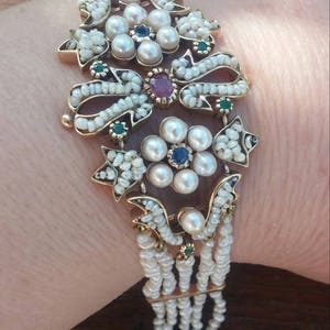 K&ouml;nnte beinhalten: Ein Armband aus Gold und Perlen mit einem floralen Design. Das Armband verf&uuml;gt &uuml;ber einen Cluster aus Perlen und Edelsteinen, darunter rote, blaue und gr&uuml;ne Steine. Das Armband besteht aus mehreren Perlenstr&auml;ngen.