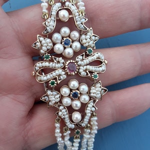K&ouml;nnte beinhalten: Ein goldenes Armband mit einem Perl- und Edelstein-Design. Das Armband verf&uuml;gt &uuml;ber einen zentralen Perlenschmuck mit einem Rubin-, Saphir- und Smaragd-Edelstein. Das Armband besteht aus mehreren Perlenschn&uuml;ren.