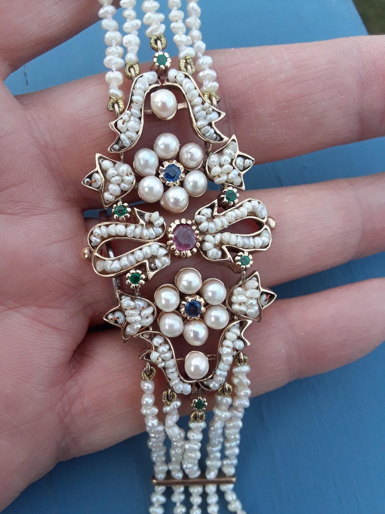 K&ouml;nnte beinhalten: Ein goldenes Armband mit einem Perl- und Edelstein-Design. Das Armband verf&uuml;gt &uuml;ber einen zentralen Perlenschmuck mit einem Rubin-, Saphir- und Smaragd-Edelstein. Das Armband besteht aus mehreren Perlenschn&uuml;ren.