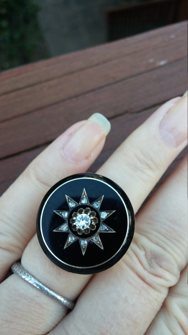 K&ouml;nnte beinhalten: Ein schwarzer Ring mit einem silbernen Sternendesign und einem klaren Strassstein in der Mitte. Der Ring befindet sich an einem Finger.