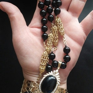 May include: A gold chain necklace with black beads and a large black oval pendant set with diamonds. The necklace is worn on a person's wrist.