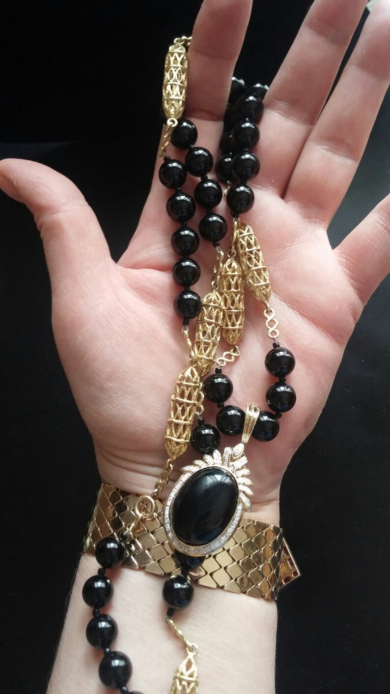 May include: A gold chain necklace with black beads and a large black oval pendant set with diamonds. The necklace is worn on a person's wrist.